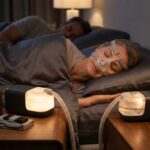 Person sleeping comfortably with a fitted nasal CPAP mask in a cozy bedroom, CPAP machine with heated tubing on the bedside table, and a travel case with accessories nearby